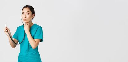 Covid-19, healthcare workers and preventing virus concept. Intrigued asian female doctor, physician in scrubs, looking away thoughtful, listening to patient lungs with stethoscope, white background photo