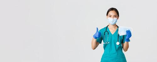 Covid-19, coronavirus disease, healthcare workers concept. Smiling asian doctor, nurse in scrubs and rubber gloves, showing thumbs-up and medical masks, white background photo