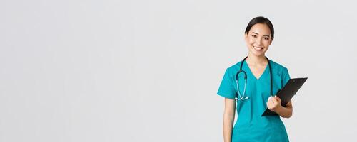 Healthcare workers, preventing virus, quarantine campaign concept. Smiling asian female nurse, doctor with clipboard wearing scrubs, provide checkup, examine patient in clinic, white background photo