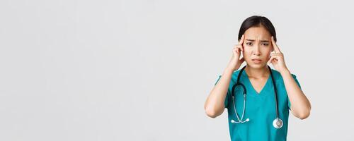 Healthcare workers, preventing virus, quarantine campaign concept. Exhausted asian female doctor feeling dizzy, touching head and grimacing from headache, standing white background photo