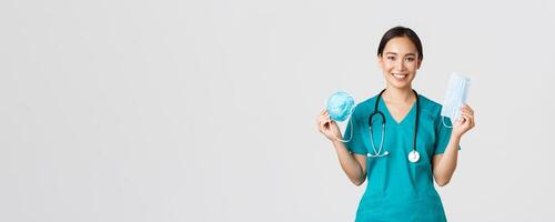 Covid-19, coronavirus disease, healthcare workers concept. Smiling pretty asian female nurse, physician in scrubs showing respirator and medical mask, suggest patients ways of virus protection photo