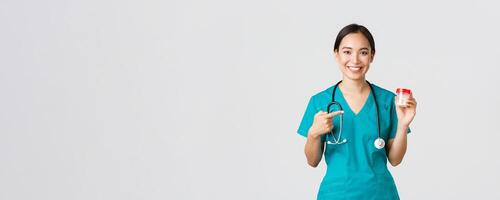Healthcare workers, preventing virus, quarantine campaign concept. Smiling pretty asian female physician, nurse in scrubs show vitamins and pointing at pills, recommend medication, white background photo