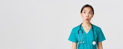 Healthcare workers, preventing virus, quarantine campaign concept. Impressed and amazed asian female doctor, nurse in medical scrubs looking upper left corner pleased, white background photo
