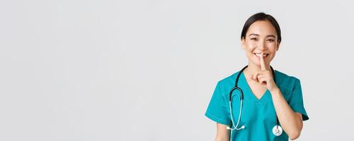 Healthcare workers, preventing virus, quarantine campaign concept. Smiling pretty asian doctor, nurse in scrubs smiling, shushing, asking stay quiet, standing white background photo