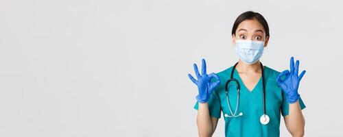 Covid-19, coronavirus disease, healthcare workers concept. Excited and impressed asian doctor, nurse in medical mask and rubber gloves showing okay gesture, approve or praise nice work photo