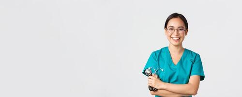 Covid-19, coronavirus disease, healthcare workers concept. Confident happy asian female physician, doctor in glasses, cross arms chest and smiling, holding stethoscope for examination photo