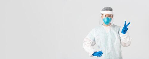 Covid-19, coronavirus disease, healthcare workers concept. Sassy asian female doctor, physician or tech lab in personal protective equipment showing peace sign, standing white background photo