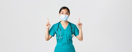 Covid-19, coronavirus disease, healthcare workers concept. Intrigued and excited asian woman doctor, nurse in medical mask and gloves, looking and pointing fingers up, white background photo