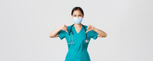 Covid-19, coronavirus disease, healthcare workers concept. Disappointed and upset asian female doctor in scrubs and medical mask showing thumbs-up, disagree or prohibit something, give advice photo