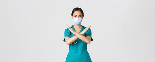 Covid-19, coronavirus disease, healthcare workers concept. Serious displeased, frowning asian doctor in scrubs show cross gesture in disapproval, stop someone, prohibit action, white background photo