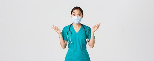 Covid-19, coronavirus disease, healthcare workers concept. Surprised and amazed asian female nurse, physician in medical mask and scrubs raising hands up excited, hear amazing news, white background photo