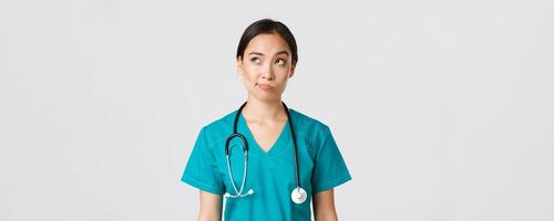 Healthcare workers, preventing virus, quarantine campaign concept. Thoughtful asian female doctor, nurse in scrubs looking upper left corner and smirk unsure, thinking, white background photo