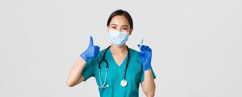 Covid-19, coronavirus disease, healthcare workers concept. Smiling beautiful asian medical worker, nurse in mask and gloves thumb-up, hold syringe with vaccine, standing white background photo