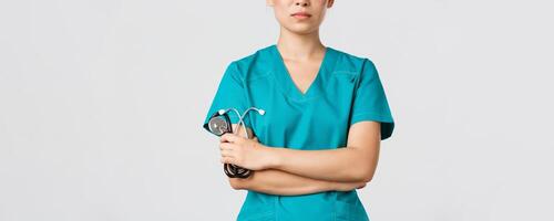Covid-19, coronavirus disease, healthcare workers concept. Close-up of confident female asian physician, doctor in medical scrubs, cross arms determined and holding stethoscope, white background photo