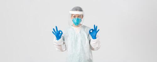 Covid-19, coronavirus disease, healthcare workers concept. Confident and serious asian female doctor, lab technician in personal protective equipment showing okay gesture in approval photo