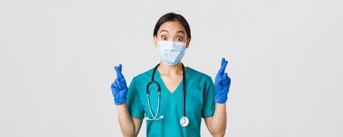 COVID-19, coronavirus enfermedad, cuidado de la salud trabajadores concepto. emocionado y esperanzado asiático hembra doctor, enfermero en médico máscara y caucho guantes cruzar dedos bueno suerte, hacer desear, blanco antecedentes foto