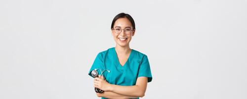 Covid-19, coronavirus disease, healthcare workers concept. Confident happy asian female physician, doctor in glasses, cross arms chest and smiling, holding stethoscope for examination photo