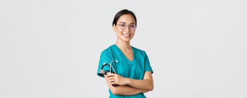 Covid-19, coronavirus disease, healthcare workers concept. Professional good-looking asian doctor, medical worker in glasses and scrubs, cross arms and smiling, white background photo