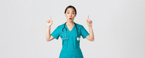 COVID-19, cuidado de la salud trabajadores, pandemia concepto. sorprendido y emocionado asiático hembra enfermero, mujer médico en matorrales preguntando pregunta interesante promoción, señalando dedos arriba, demostración clínica anuncio foto