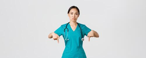 Covid-19, healthcare workers, pandemic concept. Upset and disappointed pouting asian female therapist, nurse in scrubs pointing fingers down, sulking sad, showing something with regret photo