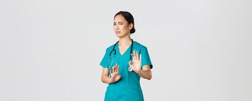 Covid-19, healthcare workers, pandemic concept. Reluctant funny asian nurse, doctor avoiding someone, step away and show stop gesture, grimacing or cringe as rejecting awful offer, refuse photo