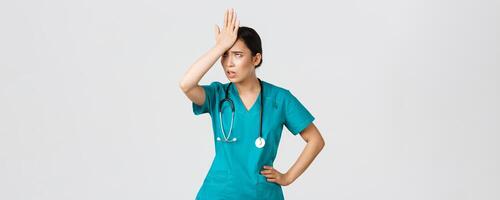 Covid-19, healthcare workers, pandemic concept. Annoyed and pissed-off asian female doctor in scrubs slap forehead, facepalm and eyeroll from someone stupid, tired standing white background photo