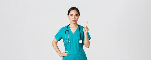 Covid-19, healthcare workers, pandemic concept. Serious-looking professional female asian doctor in scrubs, physician shaking finger in prohibition, warning gesture, forbid something photo