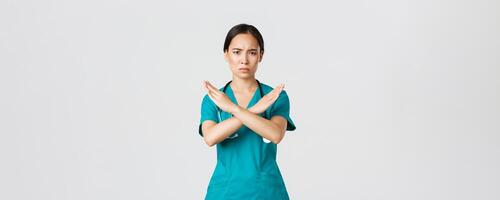 Covid-19, healthcare workers, pandemic concept. Displeased serious-looking concerned asian female doctor show cross gesture, want you stop, prohibit or forbid dangerous action, white background photo