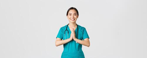 Covid-19, healthcare workers and preventing virus concept. Smiling beautiful asian female nurse, physician in scrubs smiling, hold hands together over chest in namaste, greeting gesture photo