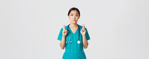 Covid-19, healthcare workers, pandemic concept. Hopeful nervous asian female intern in scrubs waiting for news, doctor cross fingers and biting lip as looking at camera, praying photo