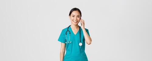 Covid-19, healthcare workers and preventing virus concept. Smiling happy asian female doctor, young intern in scrubs talking on phone, looking at camera hopeful, white background photo
