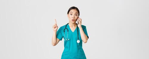 Covid-19, healthcare workers and preventing virus concept. Portrait of beautiful asian female doctor, nurse in scrubs talking on phone and raise finger as have great idea, remember, suggest plan photo