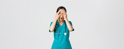 Covid-19, healthcare workers and preventing virus concept. Scared and shocked asian female nurse, doctor in scrubs witness something embarrassing or scary, cover eyes but peek through fingers photo