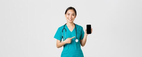 COVID-19, cuidado de la salud trabajadores y en línea medicina concepto. sonriente asiático hembra doctor, médico en matorrales señalando dedo a móvil pantalla, recomendar descargar chequeo, consulta solicitud foto