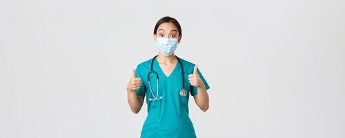Covid-19, coronavirus disease, healthcare workers concept. Impressed happy asian female doctor, physician in medical mask and scrubs show thumbs-up, agree, compliment choice, praise nice work photo