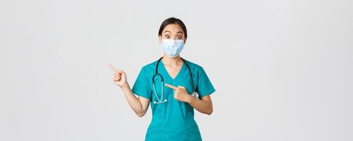 Covid-19, coronavirus disease, healthcare workers concept. Surprised and interested asian female doctor, physician in medical mask and scrubs pointing fingers upper left corner, white background photo