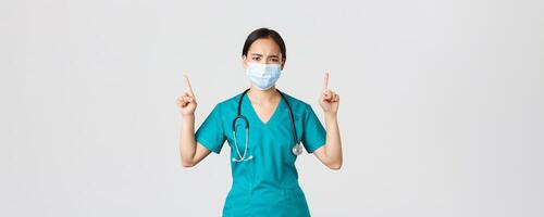 Covid-19, coronavirus disease, healthcare workers concept. Gloomy disappointed asian nurse, physician in medical mask and scrubs, pointing fingers up and frowning perplexed, look frustrated photo