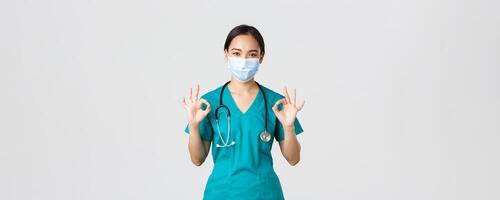 Covid-19, coronavirus disease, healthcare workers concept. Young professional female doctor, asian intern in scrubs and medical mask showing okay gesture, guarantee quality photo