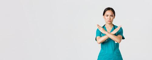 Covid-19, healthcare workers, pandemic concept. Displeased serious-looking concerned asian female doctor show cross gesture, want you stop, prohibit or forbid dangerous action, white background photo