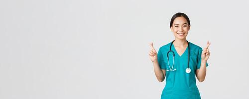 Covid-19, healthcare workers, pandemic concept. Optimistic smiling asian female doctor, physician in scrubs having faith, cross fingers good luck, making wish, hope for better, white background photo