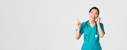 Covid-19, healthcare workers and preventing virus concept. Portrait of young pretty asian female doctor, nurse in scrubs making online order, talking on phone and pointing upper left corner, smiling photo
