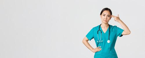 Covid-19, healthcare workers, pandemic concept. Exhausted and bothered, annoyed asian female doctor, nurse making shot gun gesture over head from boredom, white background photo