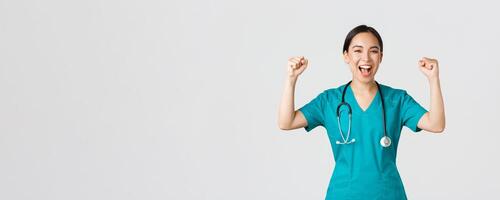 Covid-19, healthcare workers and preventing virus concept. Successful confident asian female doctor or nurse in scrubs fist pump and shouting yes, rejoicing, encourage herself, aim for victory photo