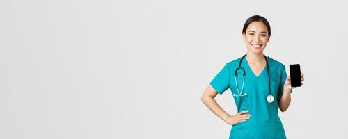 Covid-19, healthcare workers and online medicine concept. Professional female asian doctor, nurse in scrubs showing smartphone screen and smiling, showing internet appointment app, white background photo