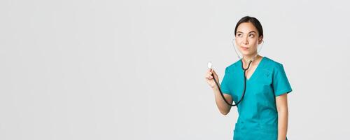 Covid-19, healthcare workers and preventing virus concept. Young focused asian female doctor, physician in scrubs, examine patient lungs, using stethoscope, standing thoughtful white background photo