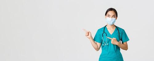 Covid-19, coronavirus disease, healthcare workers concept. Curious pleased asian female doctor, physician in medical mask and gloves, looking and pointing upper left corner with intrigued smile photo