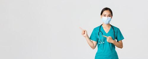 Covid-19, coronavirus disease, healthcare workers concept. Professional confident asian female physician, doctor in medical mask and scrubs pointing fingers left, showing advertisement photo