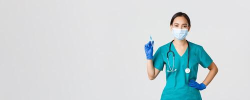 Covid-19, coronavirus disease, healthcare workers concept. Confident asian female nurse, doctor in medical mask and rubber gloves, holding syringe with vaccine, standing white background photo