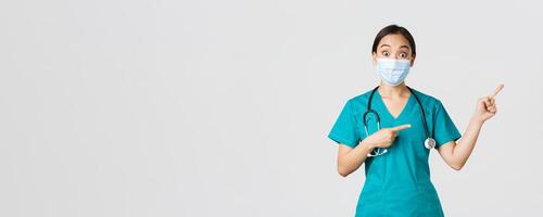 Covid-19, coronavirus disease, healthcare workers concept. Enthusiastic happy asian female doctor, physician or nurse in medical mask and scrubs, pointing upper right corner, show awesome promo photo