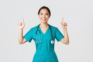 Covid-19, healthcare workers, pandemic concept. Smiling beautiful asian female nurse, doctor in scrubs pointing and looking up with satisfied grin, happy to show awesome promo offer, white background photo